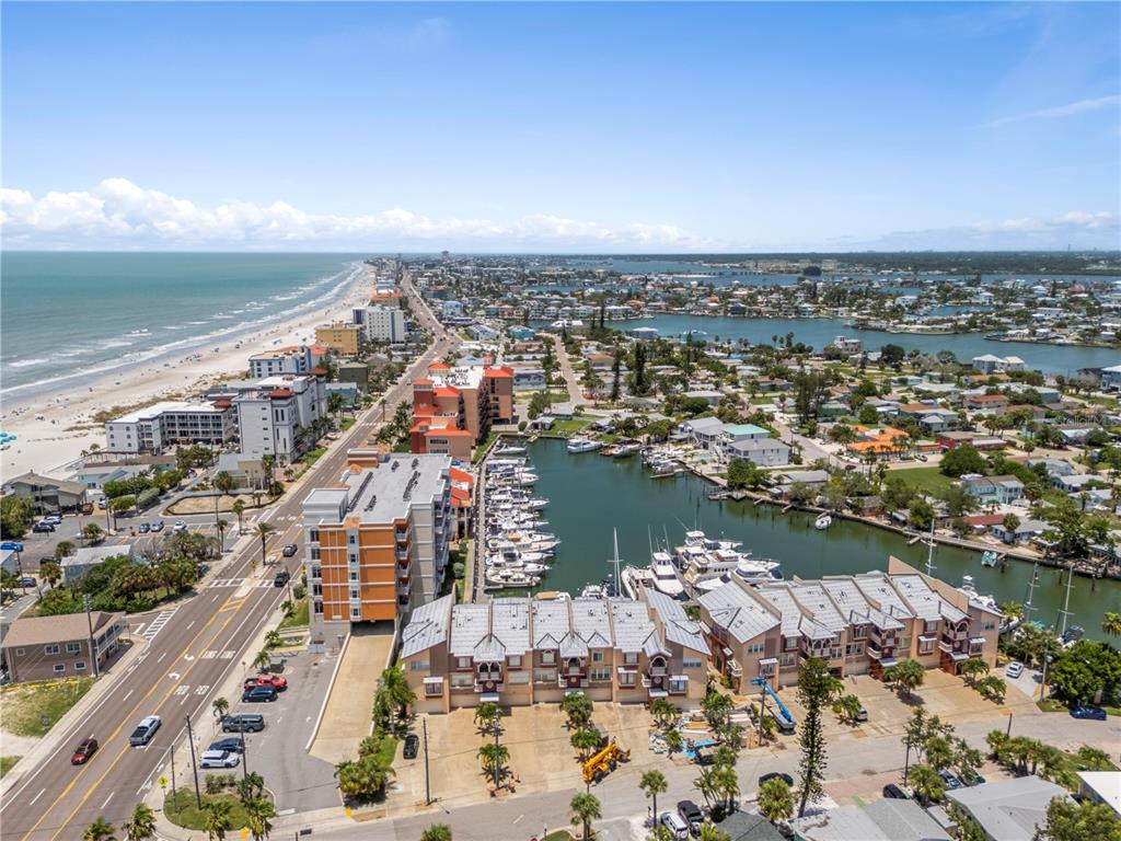 An aerial view shows a coastal town with a beach, a marina, and residential buildings — 116 131ST AVENUE E UNIT F, MADEIRA BEACH, FL