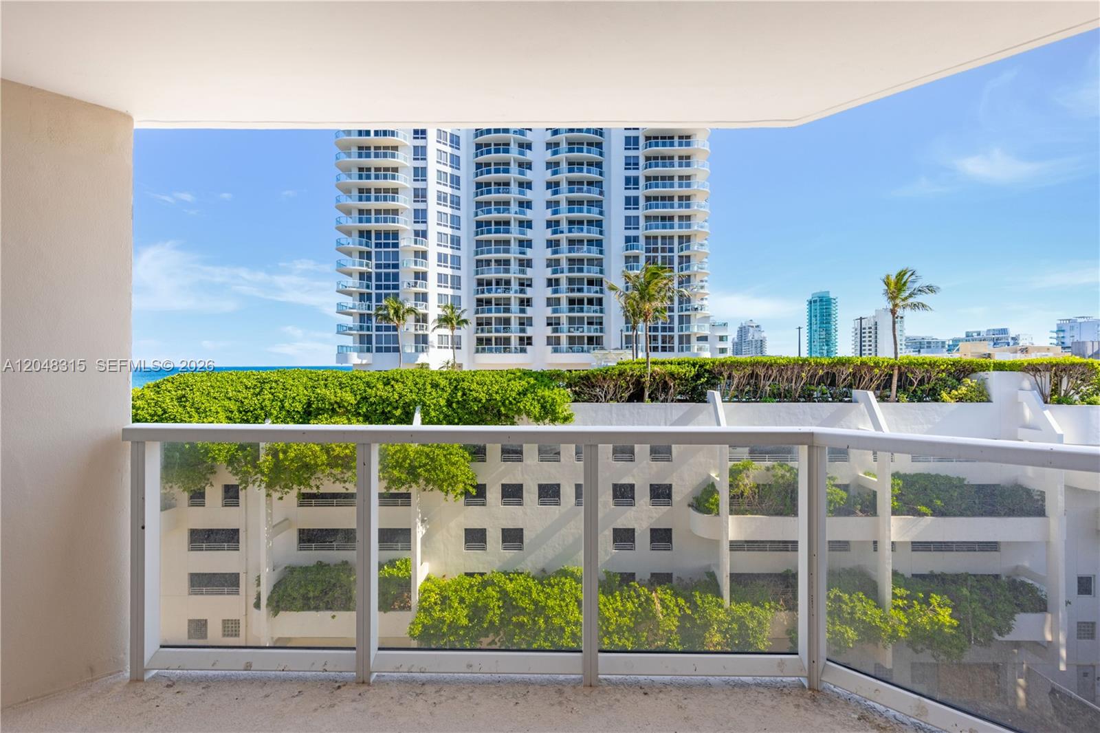 A balcony with a glass railing overlooks a parking structure, lush greenery, and high-rise buildings — 6423 Collins Ave # 703, Miami Beach FL 33141, MIAMI BEACH, FL
