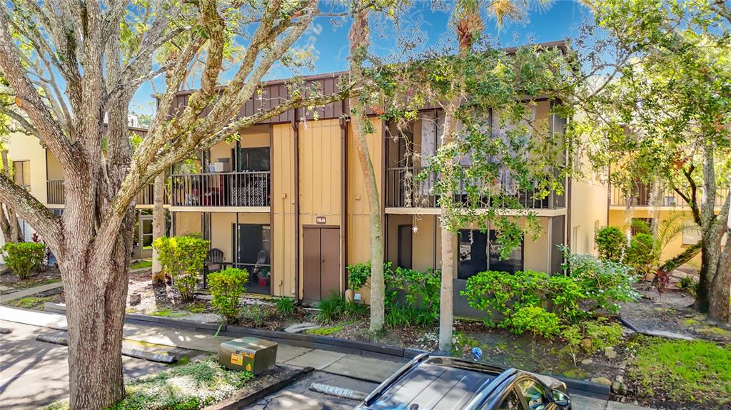 A two-story apartment building with tan siding, screened balconies, and mature trees in the foreground — 11805 RAINTREE LAKE LN UNIT D, TEMPLE TERRACE, FL