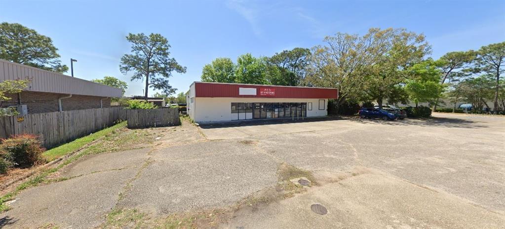 A single-story commercial building with a red roofline sits behind a large, cracked asphalt parking lot — 1167 COLLEGE BOULEVARD, PENSACOLA, FL