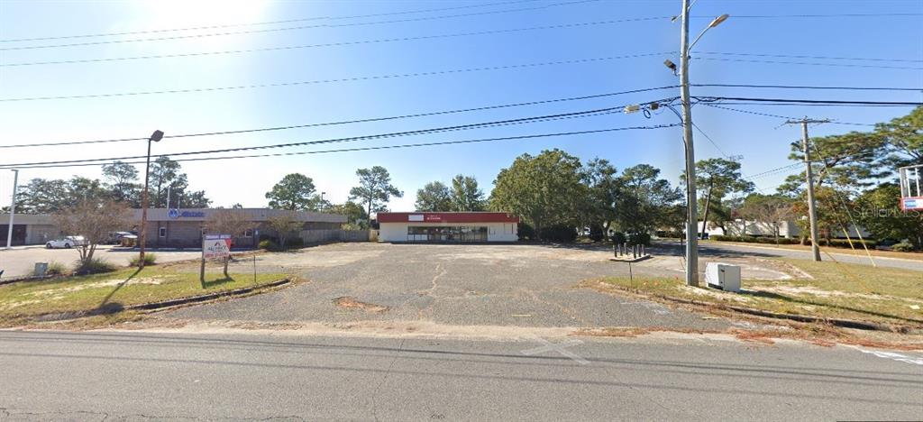 A paved commercial lot features a single-story building with a red roofline and surrounding trees — 1167 COLLEGE BOULEVARD, PENSACOLA, FL