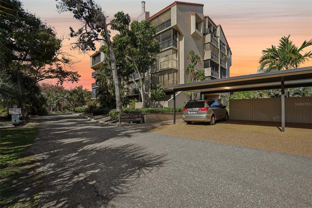 A multi-story apartment building with a covered parking area and a sign reading Sandy Cove — 4900 OCEAN BOULEVARD UNIT 201, SARASOTA, FL