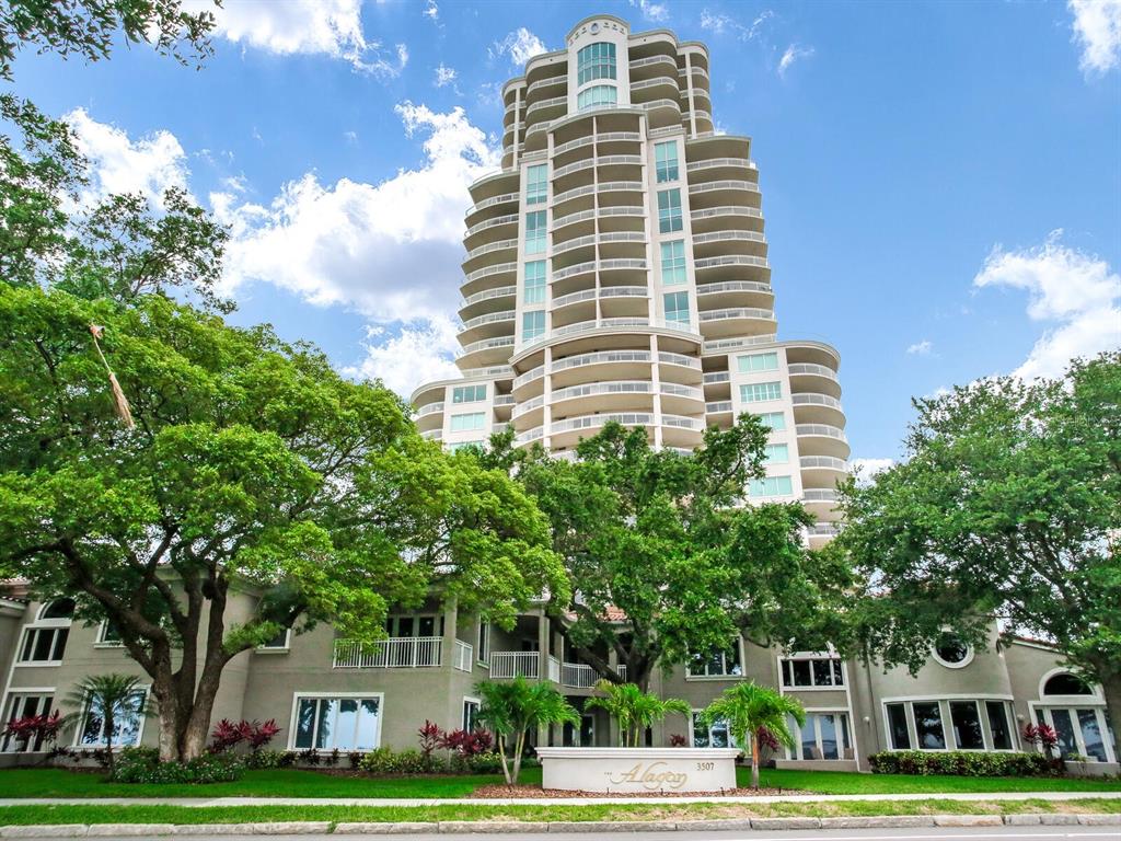 A tall, multi-story residential tower stands behind a low-rise building with lush green trees — 3507 BAYSHORE BOULEVARD UNIT 1202, TAMPA, FL