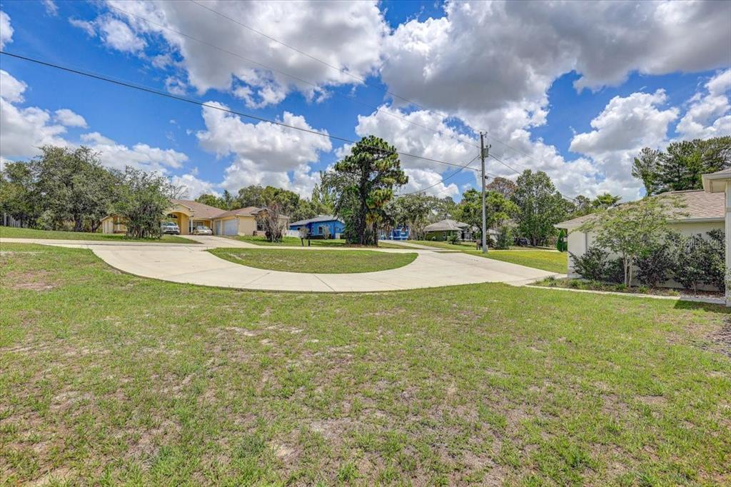 A view of a grassy front yard with a curved concrete driveway and neighboring houses — 7314 MADRID ROAD, WEEKI WACHEE, FL