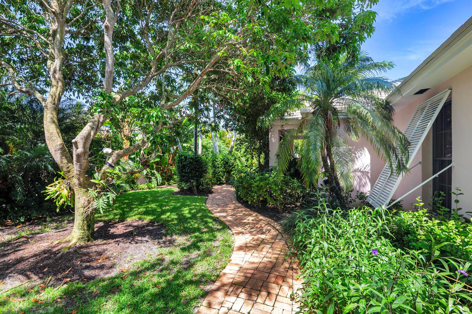 A brick pathway winds through a lush garden beside a light pink house exterior — 243 Garden Road, Palm Beach FL 33480, PALM BEACH, FL