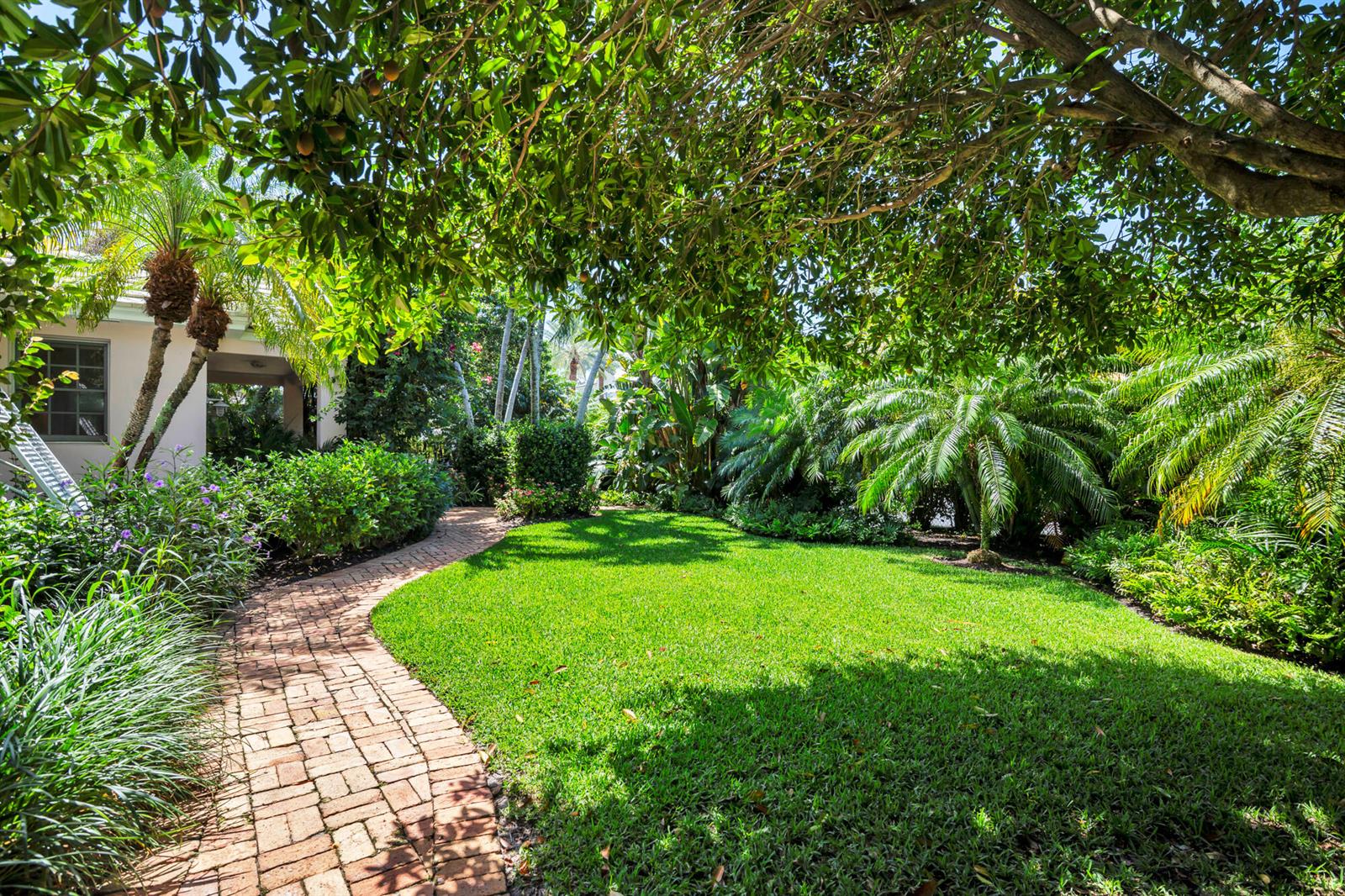 A brick path curves through a lush green lawn surrounded by tropical trees and plants — 243 Garden Road, Palm Beach FL 33480, PALM BEACH, FL