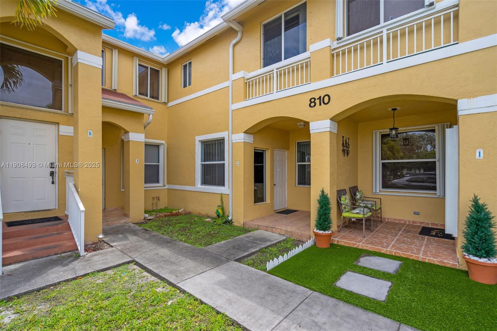 The exterior of a yellow two-story townhouse features a walkway, small patio, and landscaping — 810 NE 212th Ter # 2, Miami FL 33179, MIAMI, FL