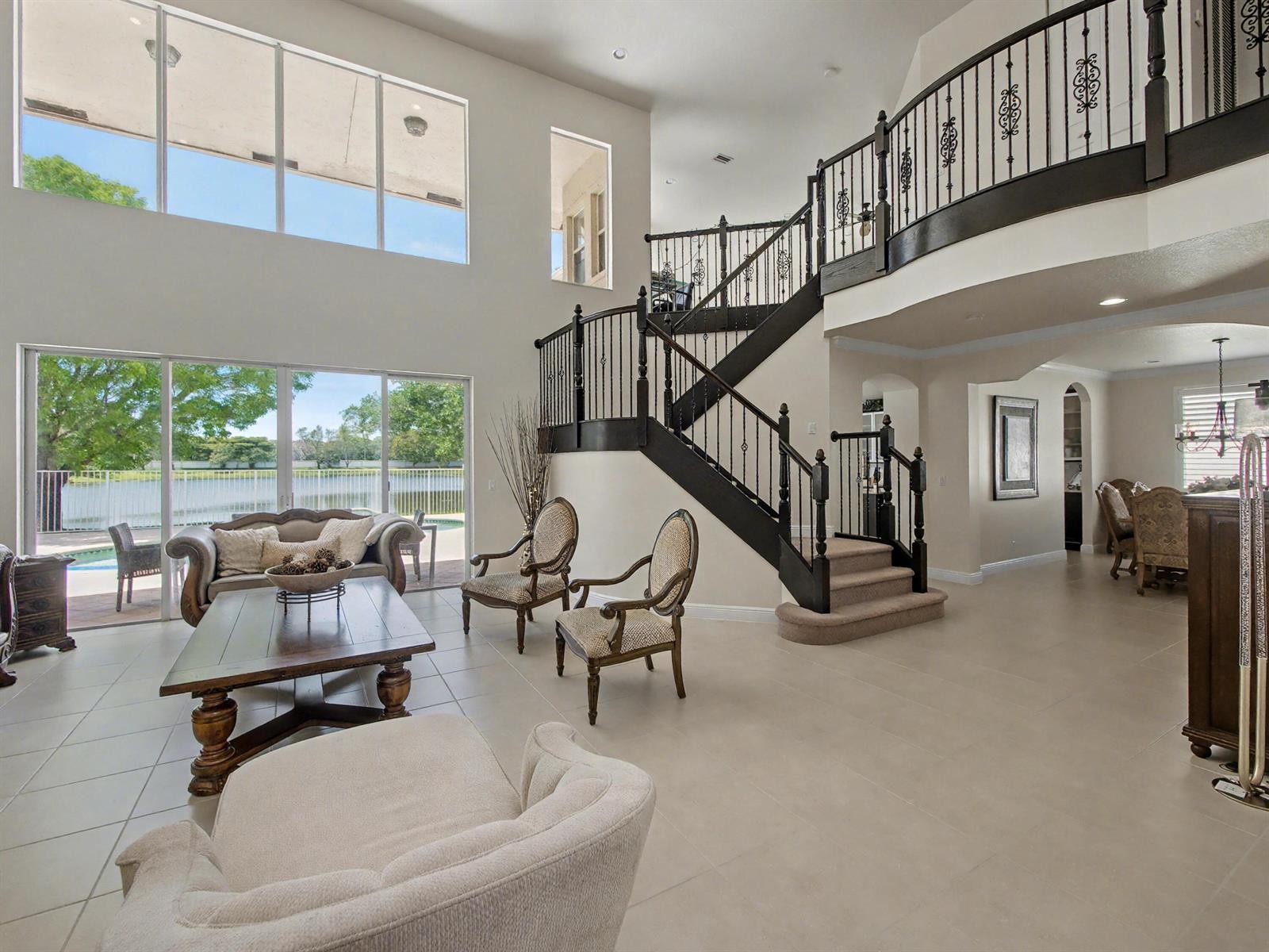 A spacious living room features a grand staircase, tile flooring, and large windows overlooking a pool — 7244 Vía Abruzzi, Lake Worth FL 33467, LAKE WORTH, FL