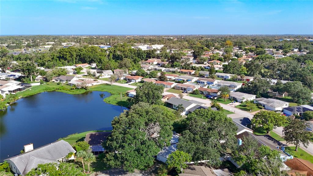 An aerial view shows a suburban neighborhood with houses surrounding a small lake — 7622 CLOVIS COURT, NEW PORT RICHEY, FL
