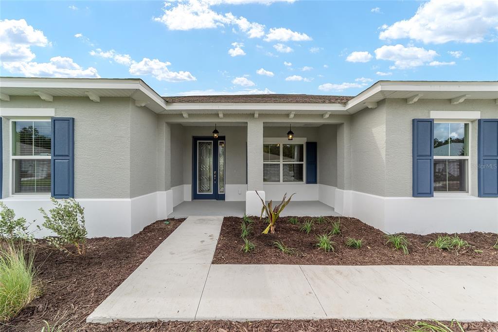 A light gray house exterior features a covered entryway, blue shutters, and a concrete walkway — 10358 SW 105TH COURT, OCALA, FL