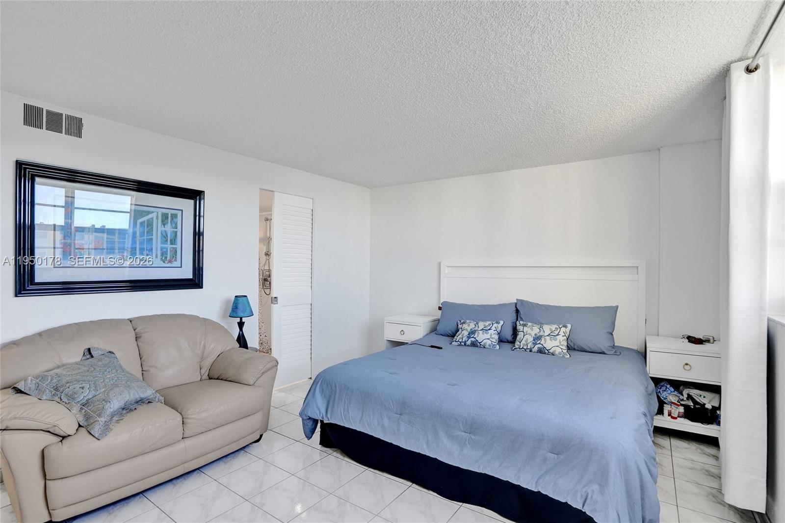 A bedroom features a bed, a beige sofa, tiled floors, and a framed wall mirror — 200 SE 5th Ave # 405, Dania Beach FL 33004, DANIA BEACH, FL