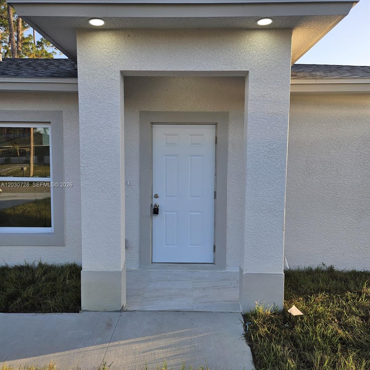 The exterior view shows a white front door recessed under a covered entryway with lighting — 3819 3rd st W, Lehigh Acres FL 33971, LEHIGH ACRES, FL