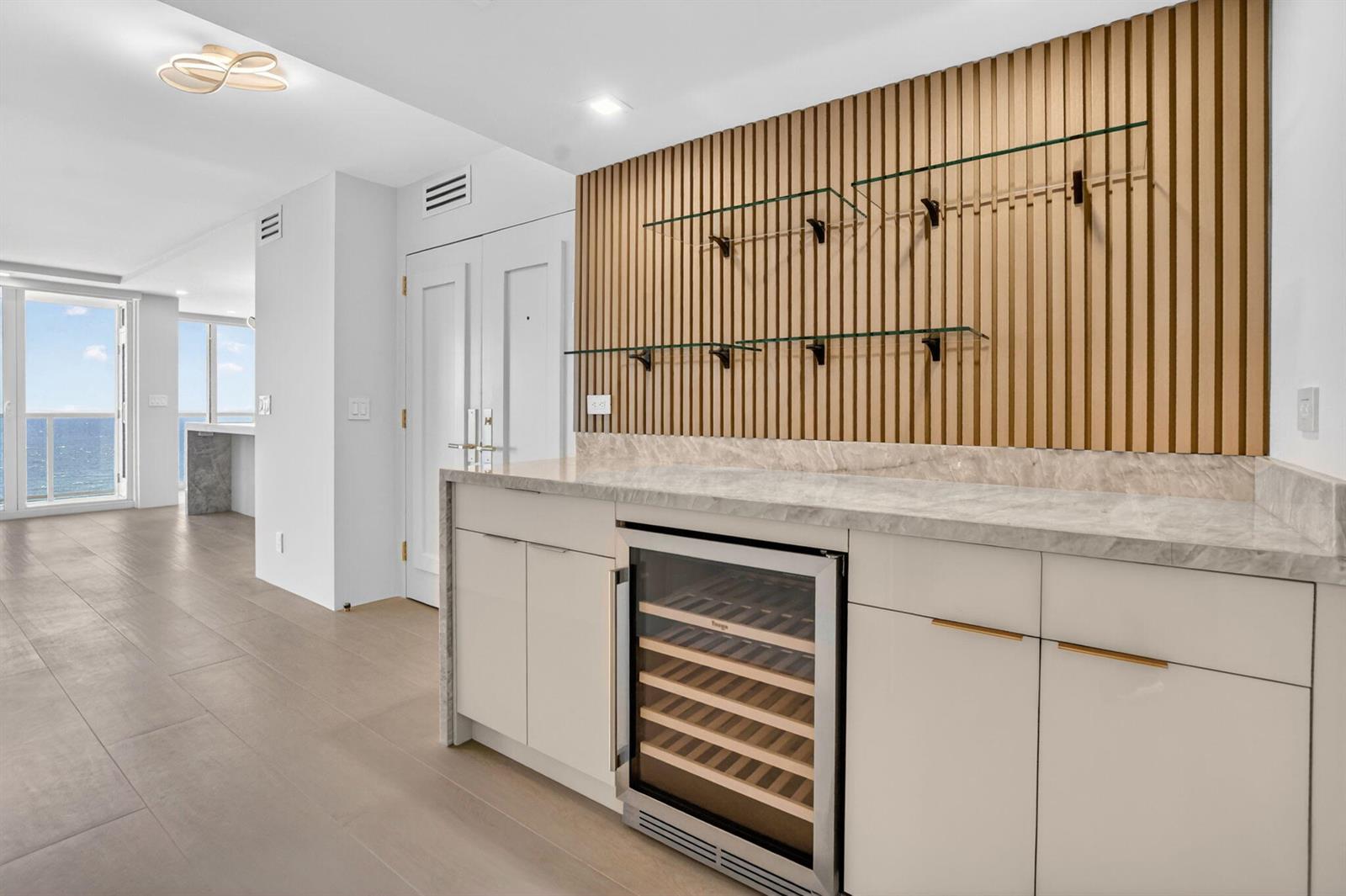 A modern wet bar features light wood slat wall paneling, glass shelves, and a wine refrigerator — 2800 S Ocean Boulevard # 23c, Boca Raton FL 33432, BOCA RATON, FL