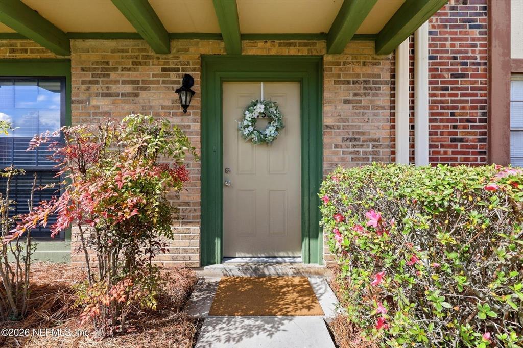A tan front door with a wreath is framed by brick walls and green trim — 8153 SUTTON PLACE N, JACKSONVILLE, FL