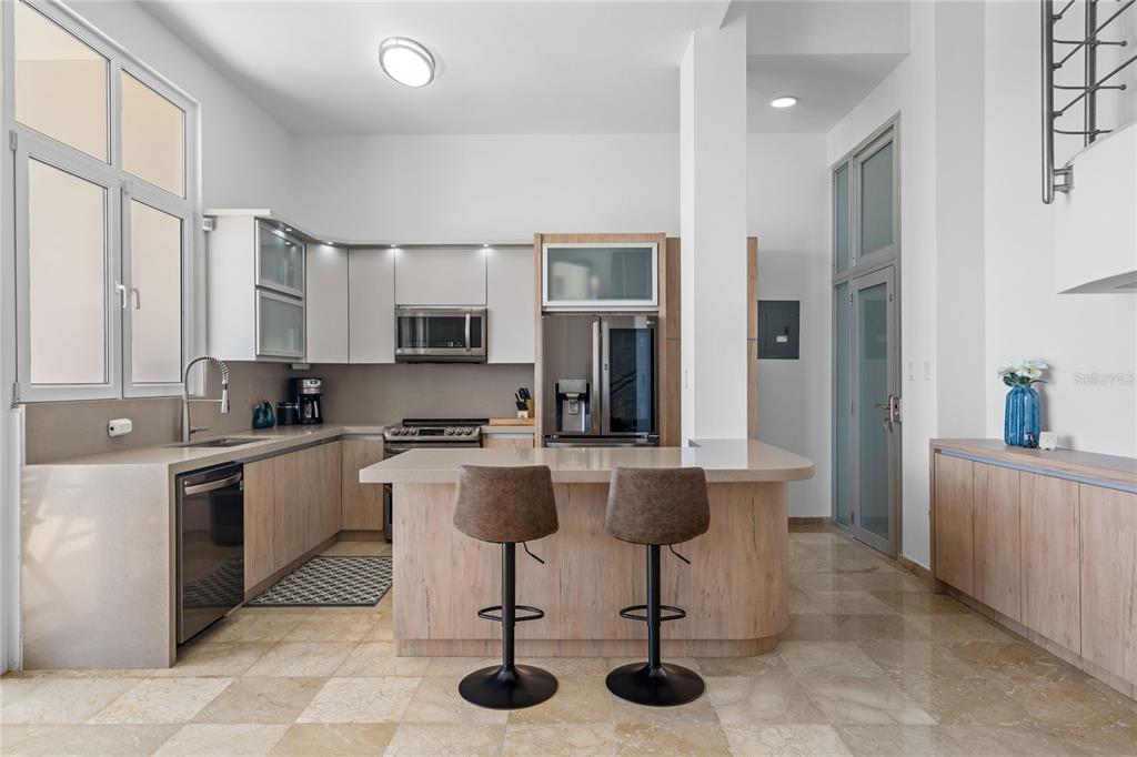 A modern kitchen features light wood cabinetry, a stone-topped island with two stools, and tile flooring — 1 SHELL CASTLE CLUB UNIT 78, HUMACAO, PR