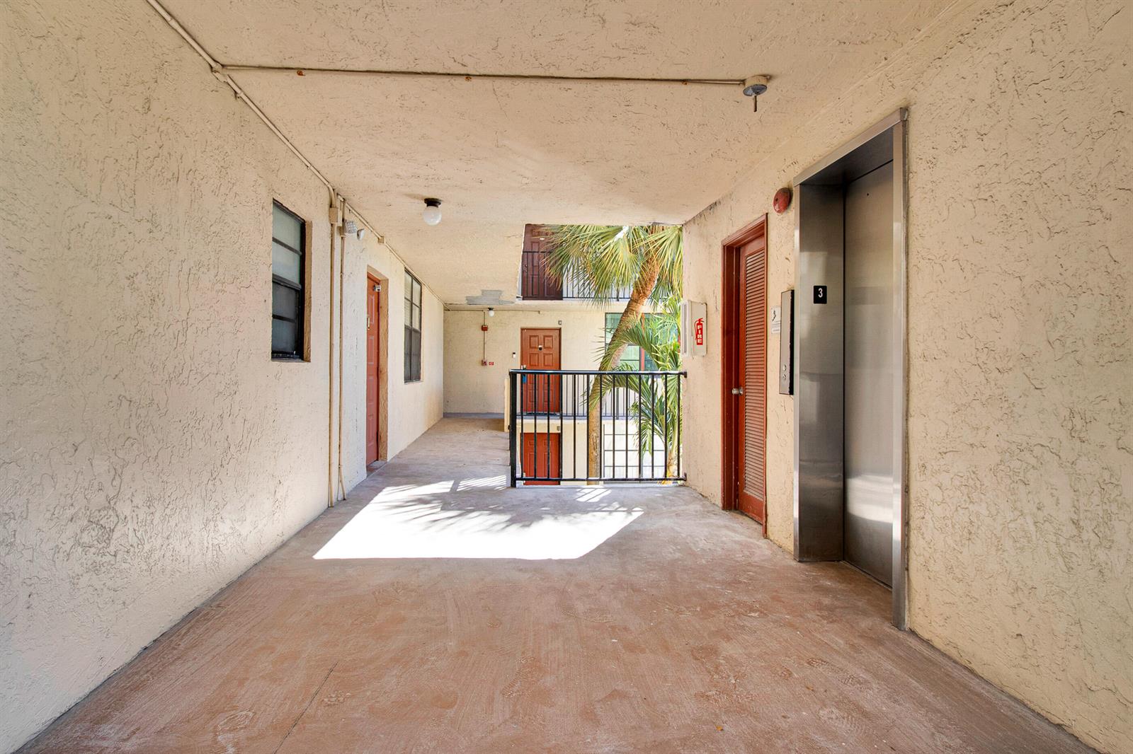 An outdoor apartment building hallway features a metal elevator, a black railing, and several doors — 3450 Blue Lake Drive # 305, Pompano Beach FL 33064, POMPANO BEACH, FL