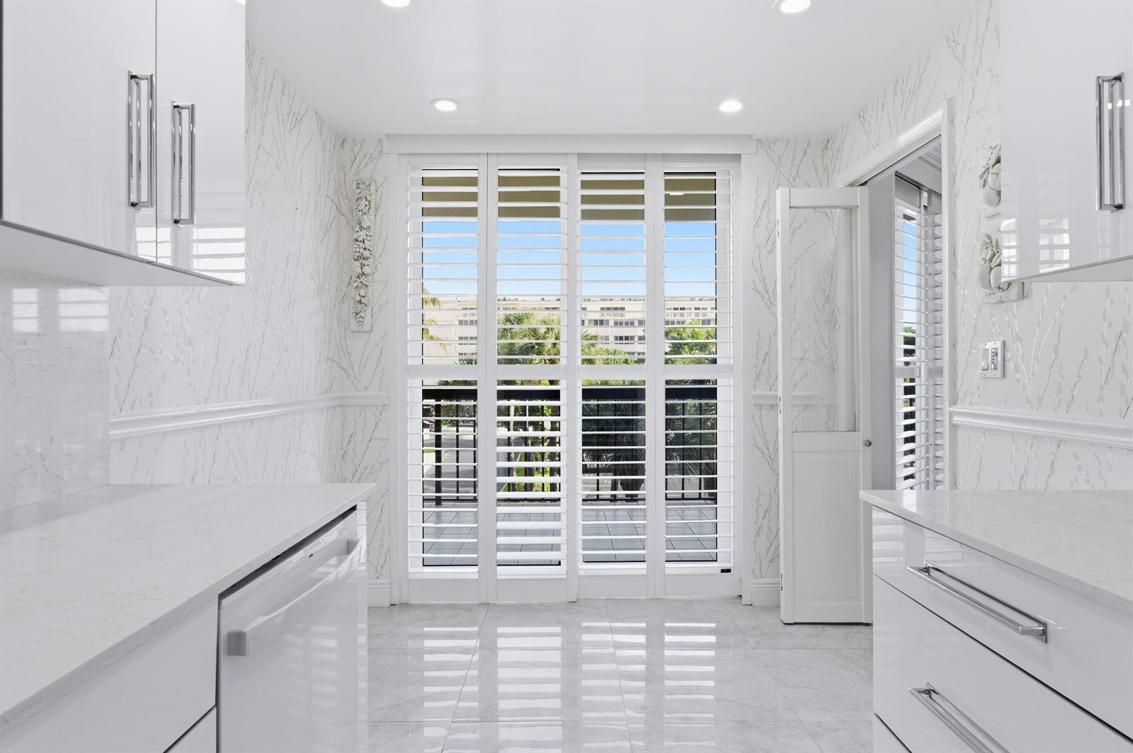 A bright kitchen with white cabinetry, marble-patterned wallpaper, and white plantation shutters over sliding doors — 2778 S Ocean Boulevard # 206s, Palm Beach FL 33480, PALM BEACH, FL