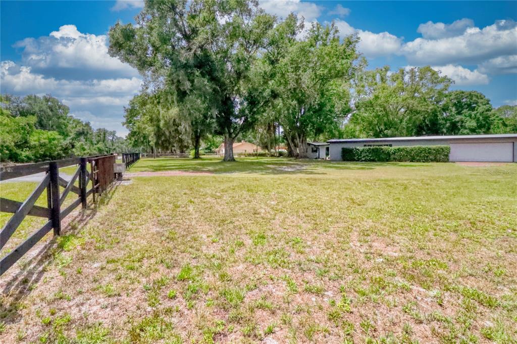A grassy yard features a black wooden fence, large trees, and a detached garage building — 5205 FIVE ACRE ROAD, PLANT CITY, FL