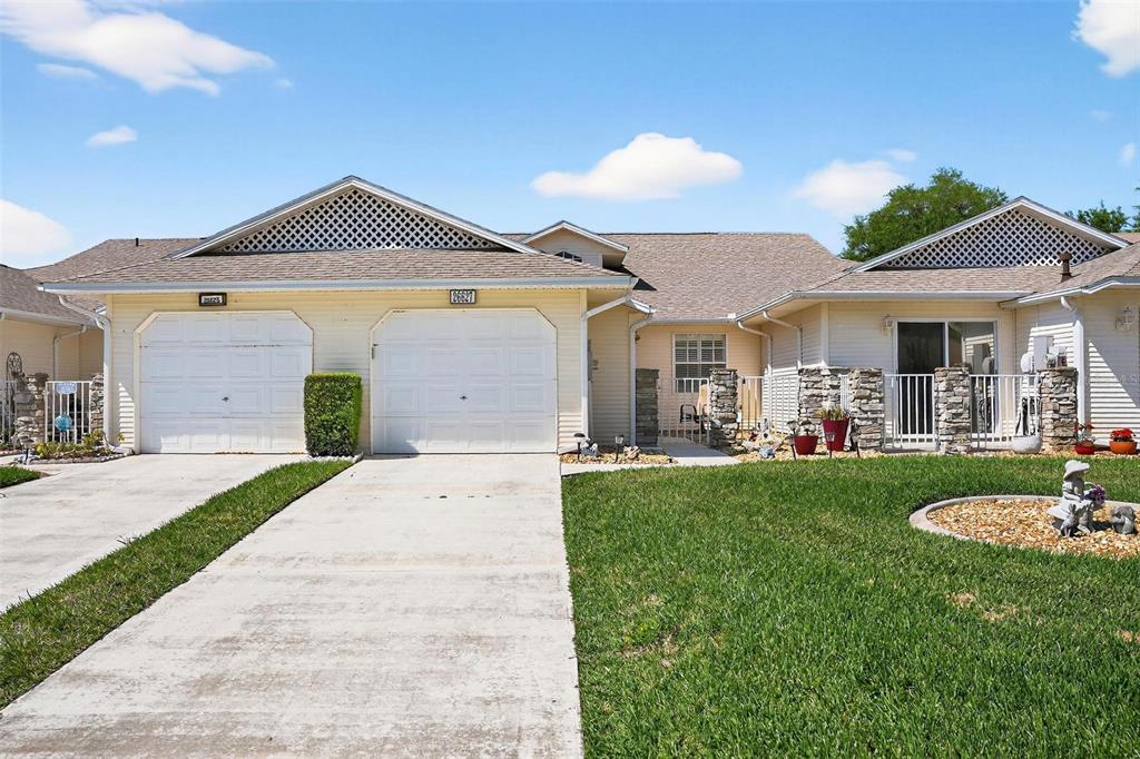 A single-story duplex with two white garage doors, light siding, and a grassy front yard — 26627 RACQUET CIRCLE, LEESBURG, FL
