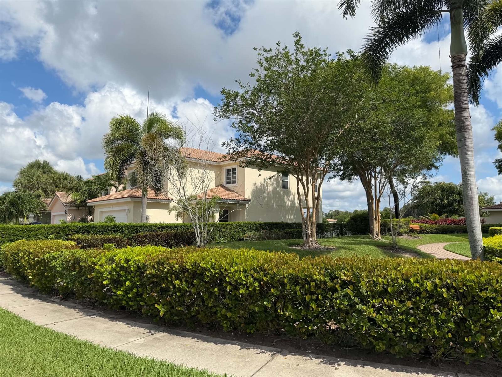 A two-story tan house with a tiled roof sits behind a manicured hedge and trees — 6355 Adriatic Way, West Palm Beach FL 33413, WEST PALM BEACH, FL