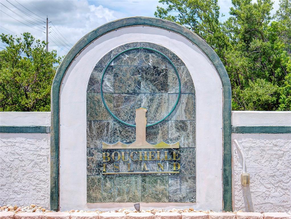 A stone monument sign for Bouchelle Island stands outdoors against a background of green trees — 444 BOUCHELLE DRIVE UNIT 201, NEW SMYRNA BEACH, FL