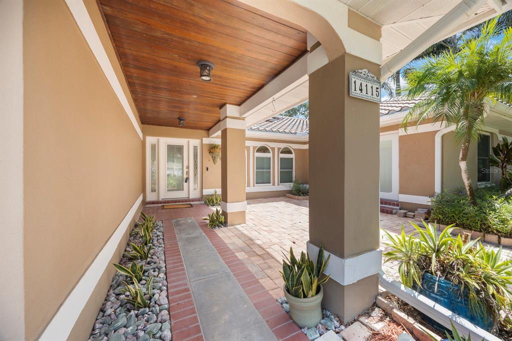 A covered entryway features a wood-paneled ceiling, brick-bordered walkway, and tan stucco walls — 14115 HARBORWOOD DRIVE, LARGO, FL