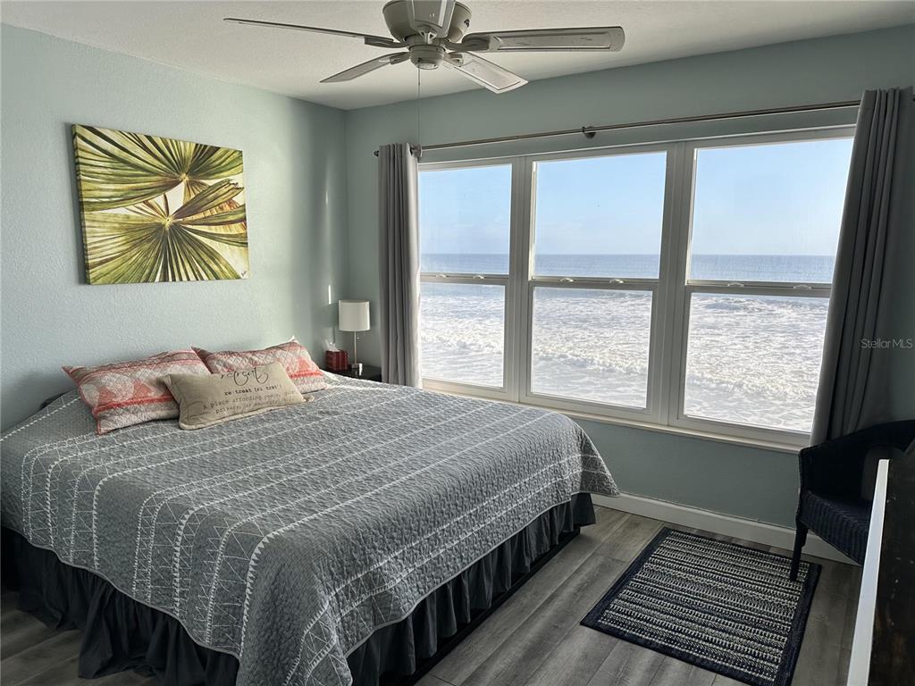 A bedroom features a bed with a gray quilt, a ceiling fan, and large ocean-view windows — 4849 SAXON DRIVE UNIT A210, NEW SMYRNA BEACH, FL