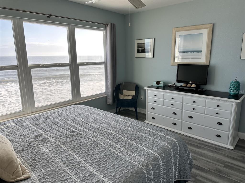 A bedroom features a white dresser, a wicker chair, and large windows overlooking the ocean — 4849 SAXON DRIVE UNIT A210, NEW SMYRNA BEACH, FL
