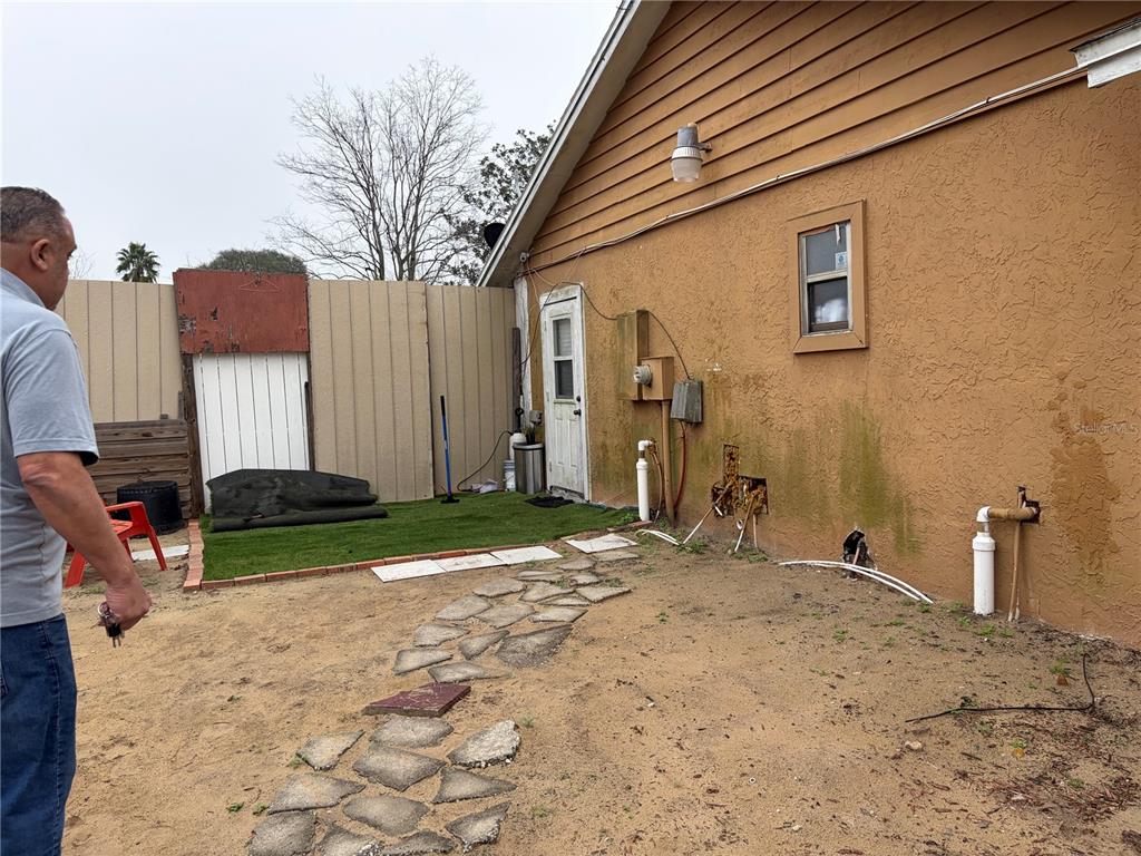 A man stands in a sandy backyard next to a tan house with exposed pipes — 1311 WOODMAN WAY, ORLANDO, FL