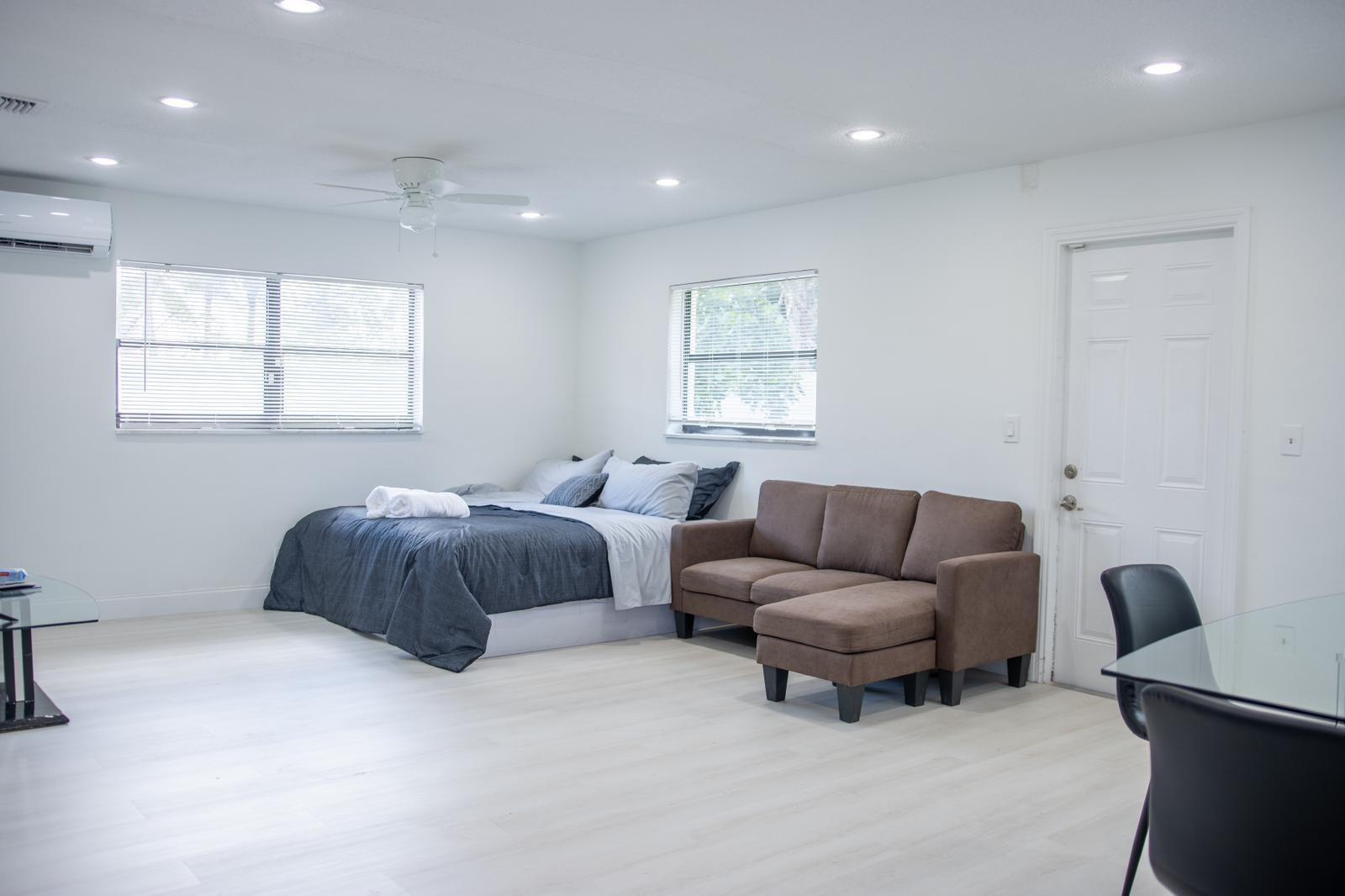 A studio apartment features white walls, light flooring, a bed, a brown sofa, and windows — 320 NW 4th Avenue # 3, Deerfield Beach FL 33441, DEERFIELD BEACH, FL