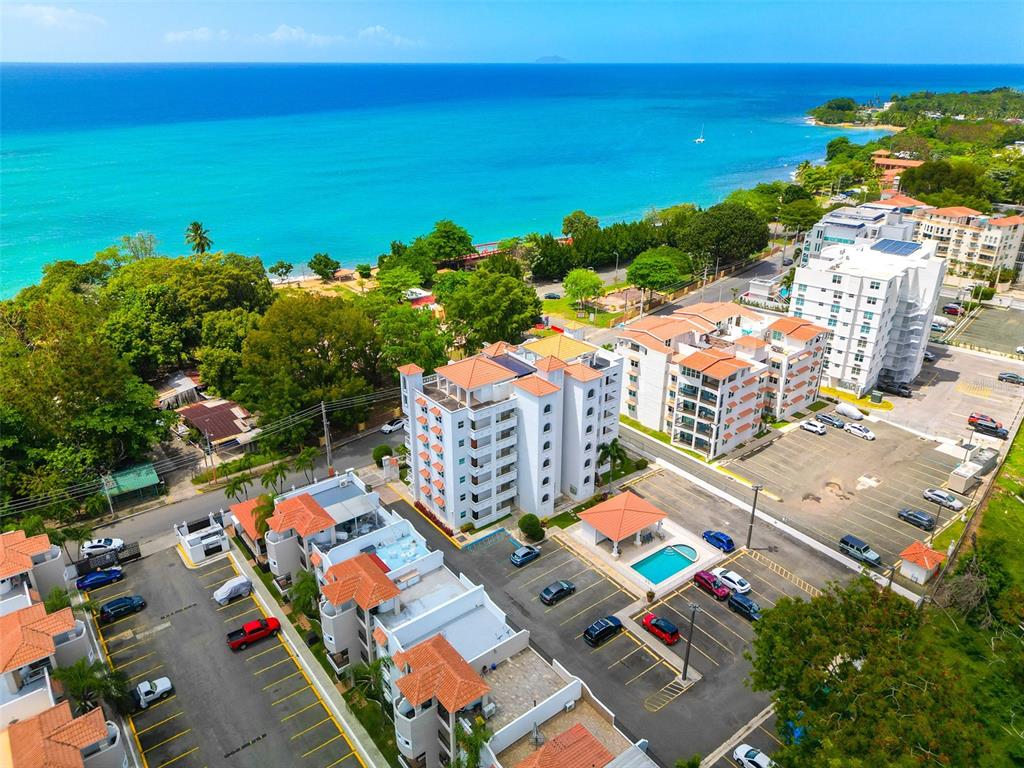 An aerial view shows a multi-story apartment complex with parking lots near a coastal beach — PR-115 ENSENADA WARD RINCON WAVE VIEW UNIT 1B, RINCON, PR