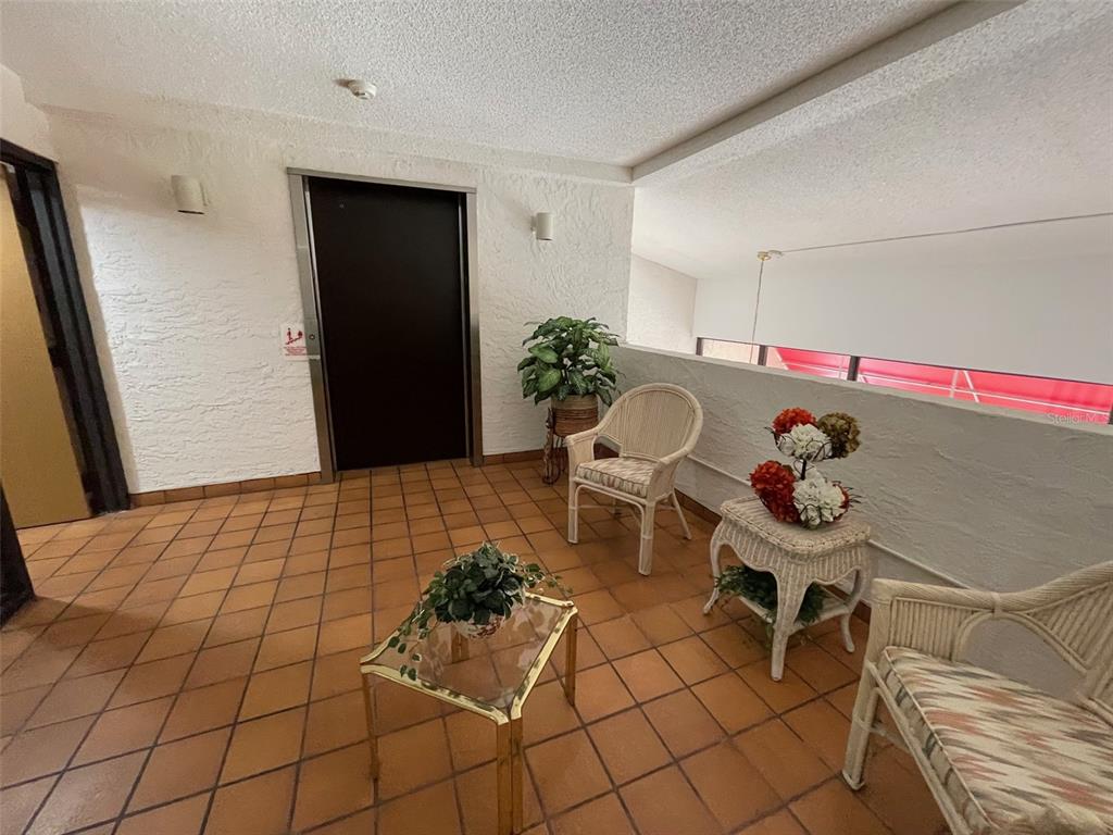 A tiled hallway features a dark elevator door, wicker furniture, and a glass-topped table — 2591 COUNTRYSIDE BOULEVARD UNIT 5203, CLEARWATER, FL