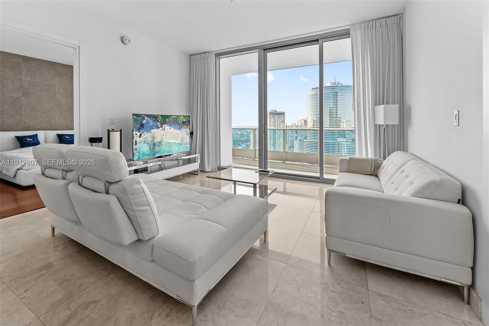 A modern living room features white leather sofas, tile flooring, and a balcony with city views — 200 Biscayne Boulevard Way # 4510, Miami FL 33131, MIAMI, FL