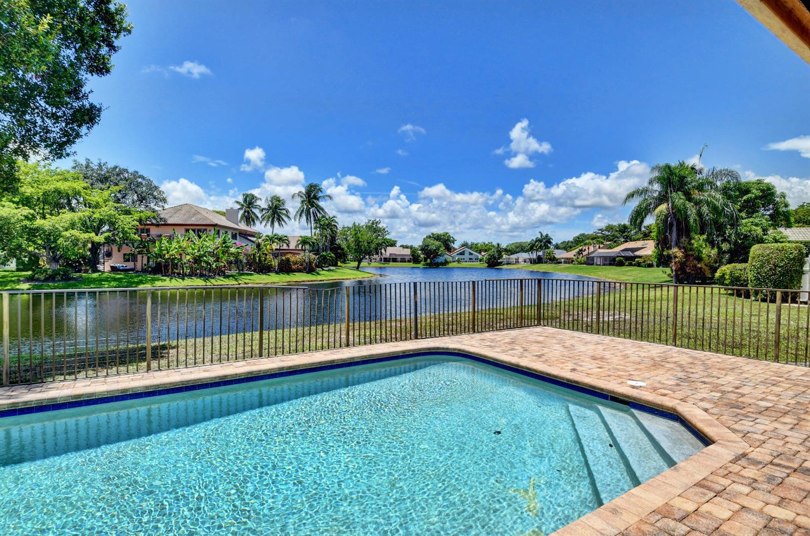 A swimming pool with a brick deck overlooks a lake and houses under a blue sky — 3110 Equestrian Drive, Boca Raton FL 33434, BOCA RATON, FL