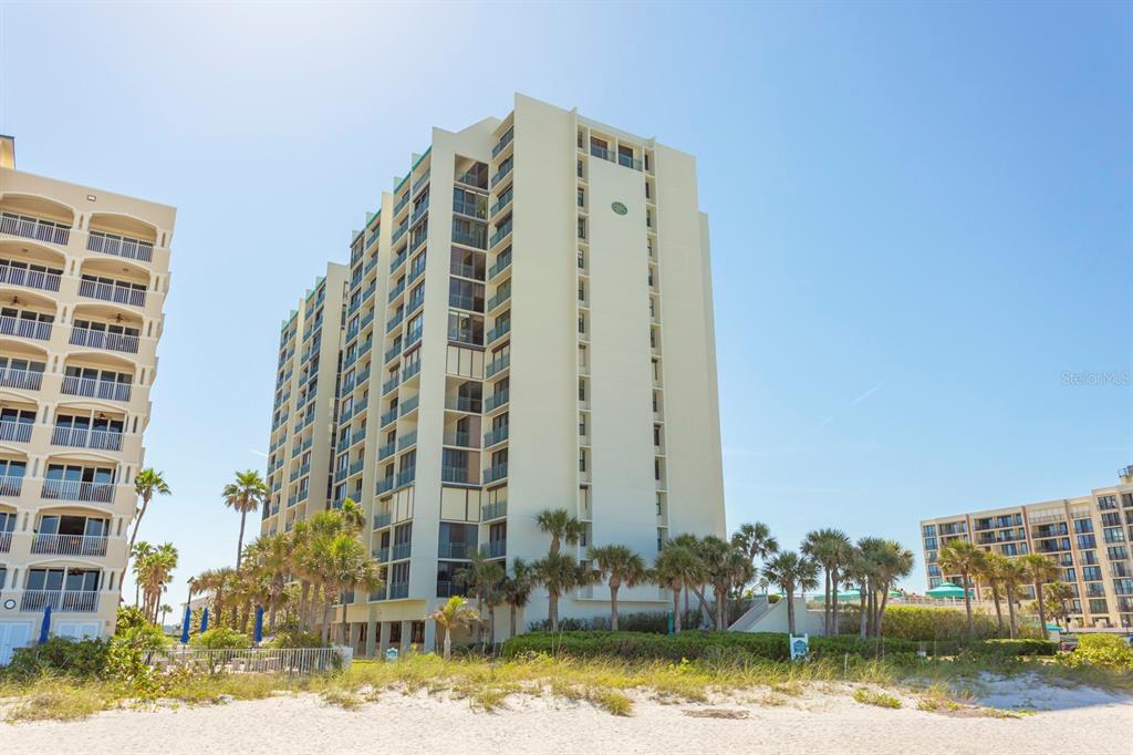 A tall, light-colored multi-story condominium building stands behind a sandy beach with palm trees — 1380 GULF BOULEVARD UNIT 206, CLEARWATER, FL