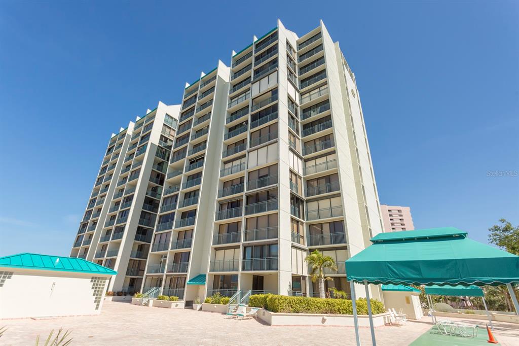 A tall, multi-story residential building with balconies stands above a paved patio area — 1380 GULF BOULEVARD UNIT 206, CLEARWATER, FL