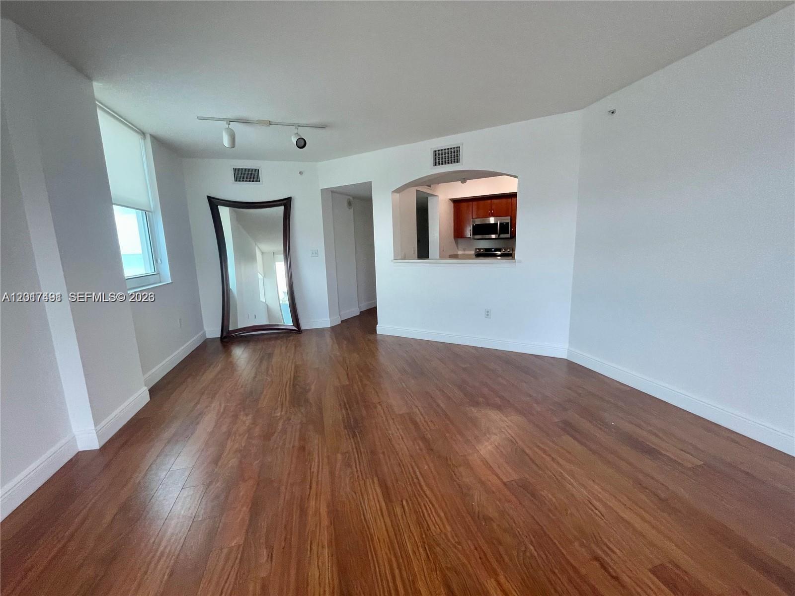 An empty room features hardwood floors, a large floor mirror, and a kitchen pass-through window — 9201 Collins Ave # 321, Surfside FL 33154, SURFSIDE, FL