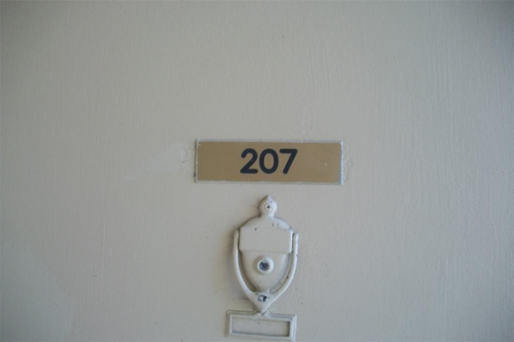 A white door features a gold-toned sign with the number 207 and a door knocker — 2616 COVE CAY DRIVE UNIT 207, CLEARWATER, FL