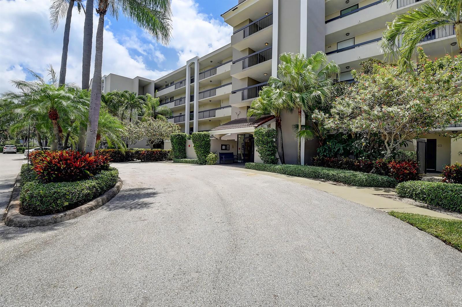 A multi-story apartment building features a paved driveway, palm trees, and landscaped garden beds — 950 Lavers Circle # F307, Delray Beach FL 33444, DELRAY BEACH, FL