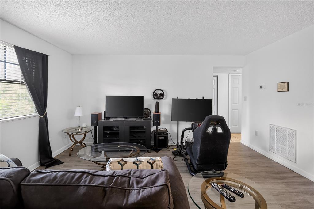 A living room features wood-look flooring, a television console, and a racing simulator setup — 12405 OAK CEDAR PLACE UNIT 202, TAMPA, FL
