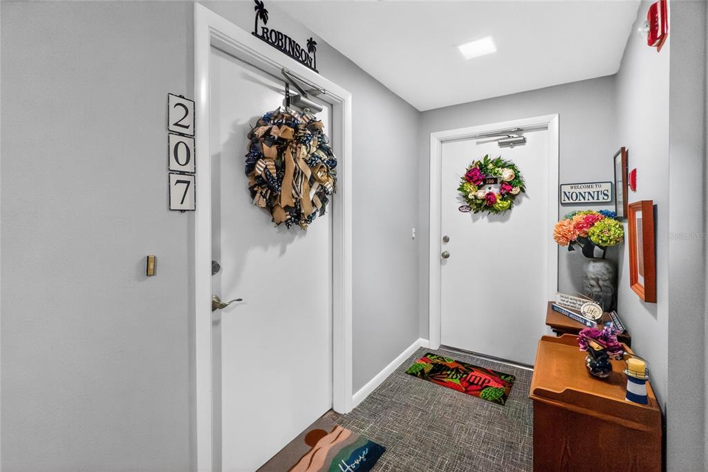 An indoor hallway shows two white doors with wreaths, wall decor, and a wooden console table — 25188 MARION AVENUE UNIT F207, PUNTA GORDA, FL
