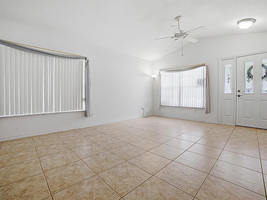 An empty living room features beige tile flooring, white walls, vertical blinds, and a ceiling fan — 25114 CRANES ROOST CIRCLE, LEESBURG, FL