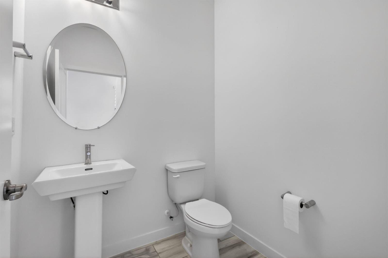 A minimalist half bathroom features a pedestal sink, round mirror, and white toilet — 8792 Antarus Drive, Lake Worth FL 33467, LAKE WORTH, FL