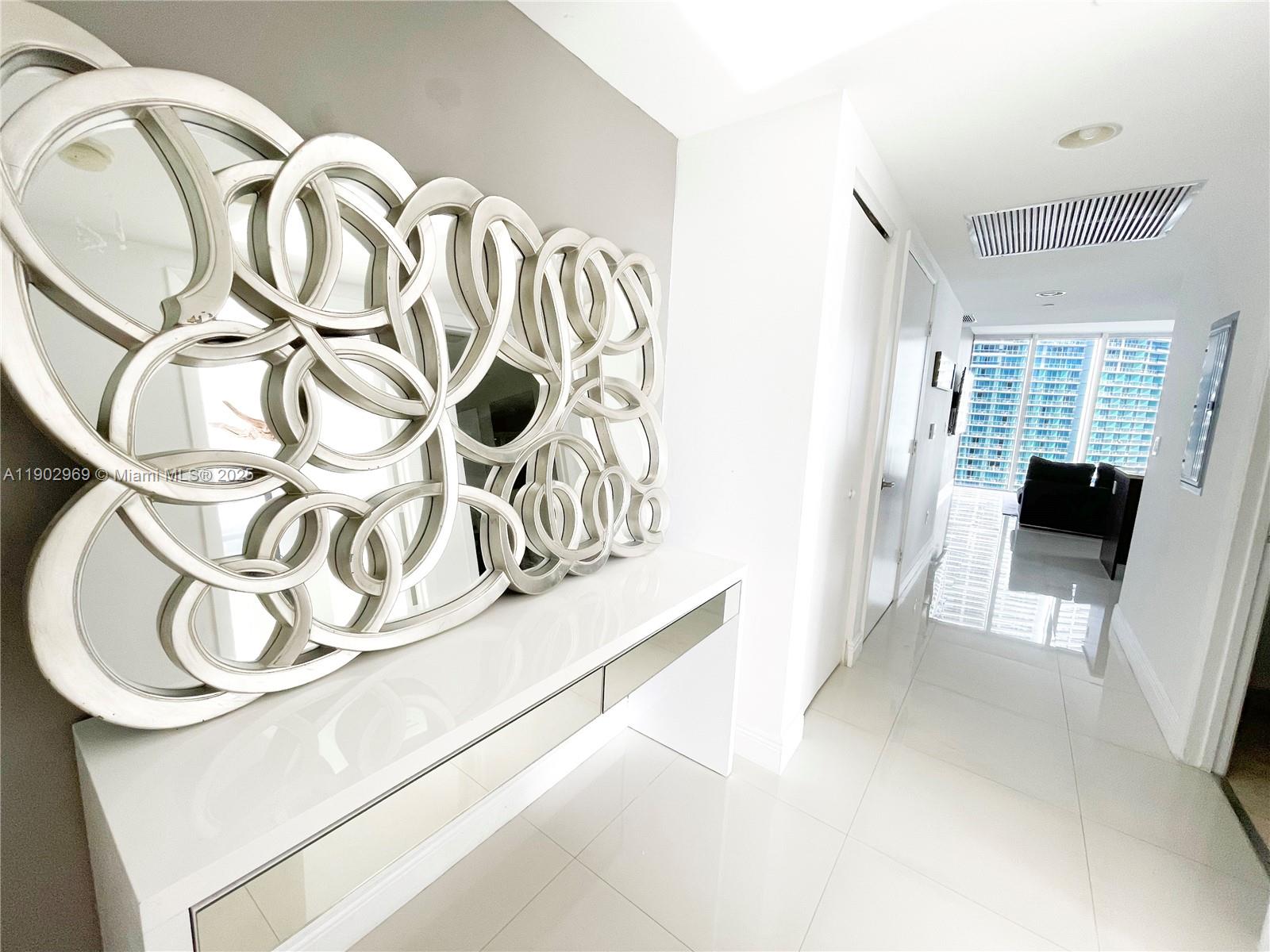 A white hallway features a large decorative circular mirror above a sleek console table — 485 Brickell Ave # 2602, Miami FL 33131, MIAMI, FL