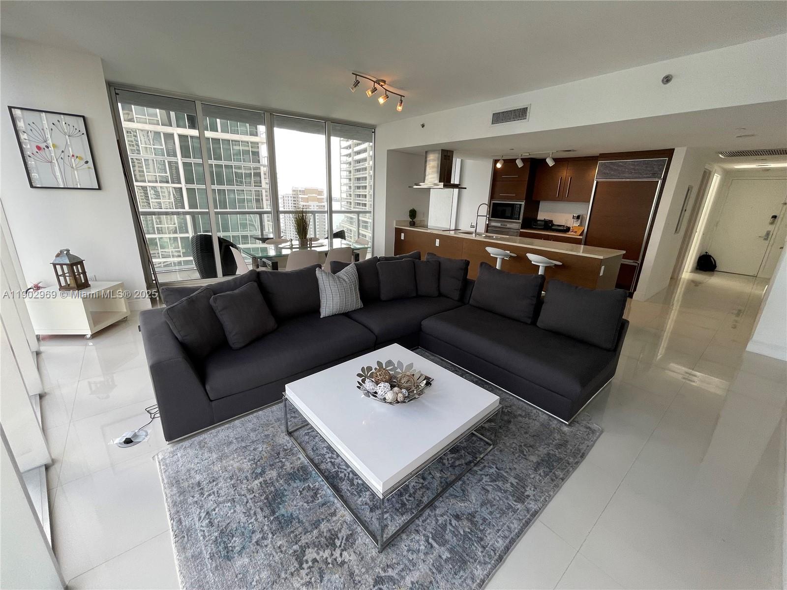 A modern living room features a dark sectional sofa, white coffee table, and open kitchen — 485 Brickell Ave # 2602, Miami FL 33131, MIAMI, FL