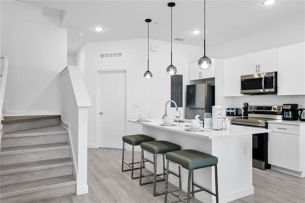 A modern kitchen features white cabinetry, a quartz island with bar stools, and adjacent stairs — 412 FAMAGUSTA DRIVE, DAVENPORT, FL