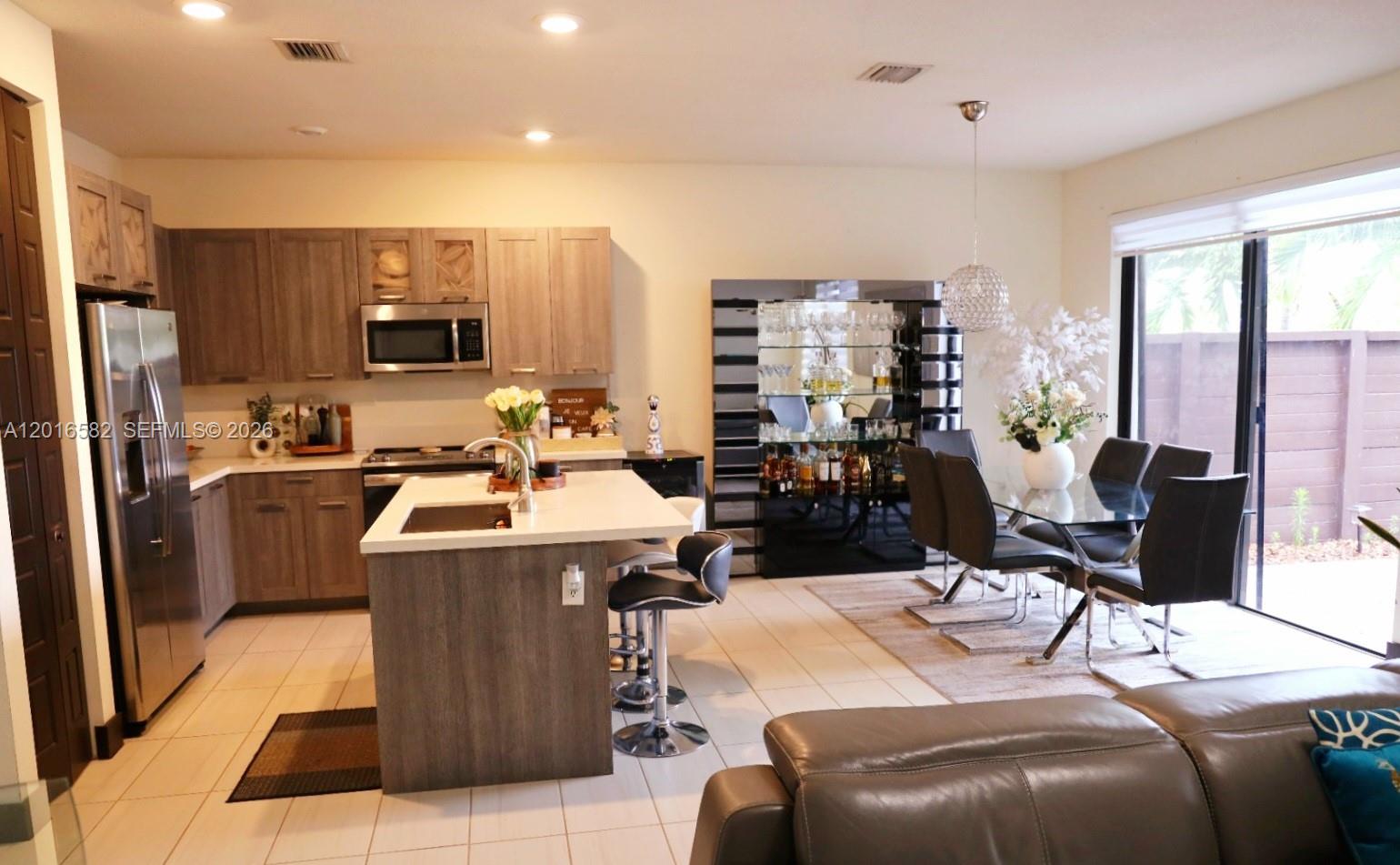 A modern kitchen features wood-toned cabinets, a central island with bar stools, and dining area — 16012 NW 91st Ct, Miami Lakes FL 33018, MIAMI LAKES, FL