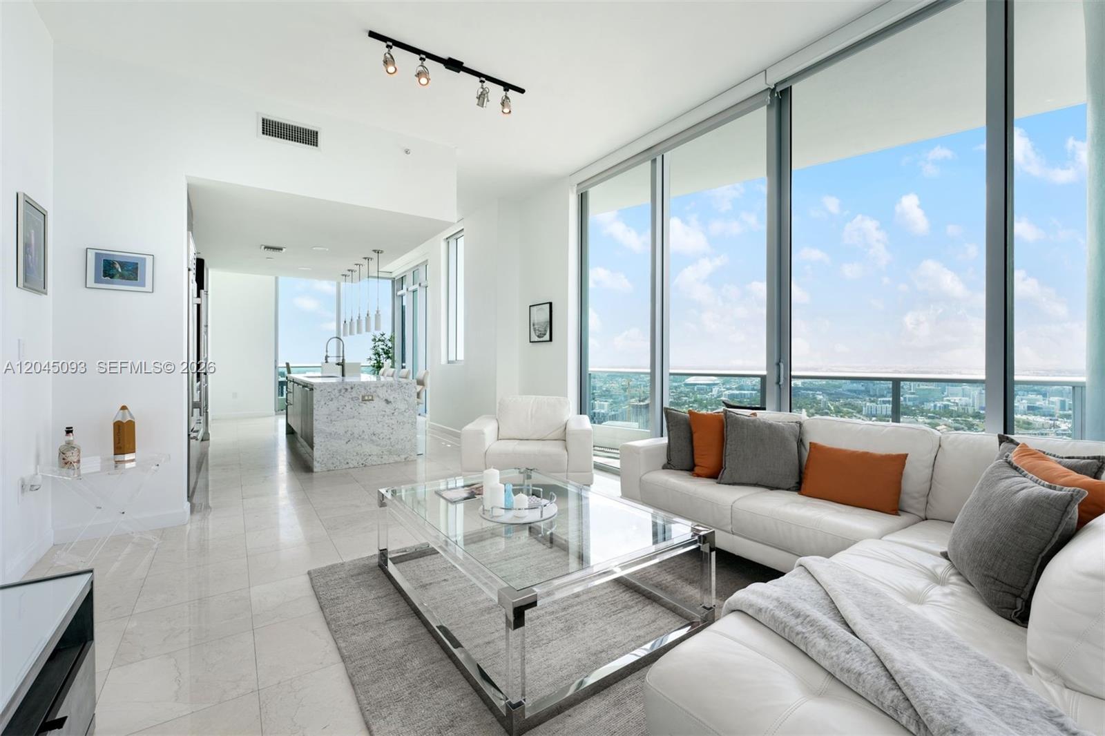 A modern living room features white leather furniture, a glass coffee table, and floor-to-ceiling windows — 900 Biscayne Blvd # 5812, Miami FL 33132, MIAMI, FL