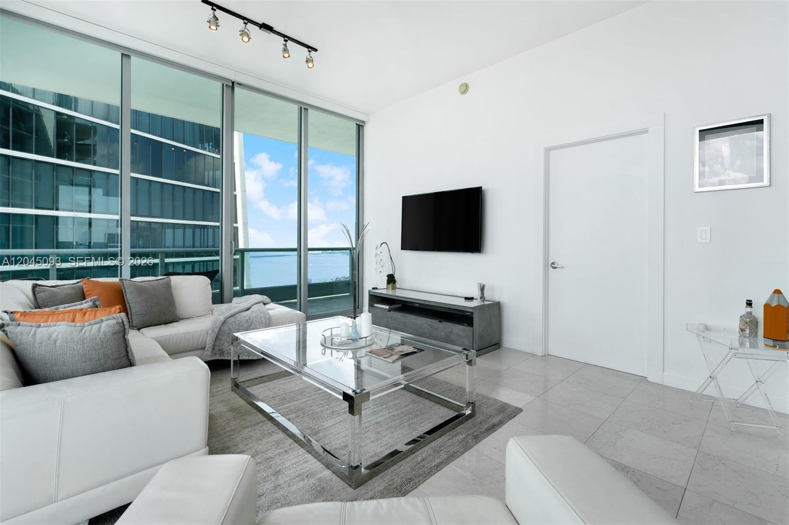 A modern living room features white leather furniture, a glass coffee table, and floor-to-ceiling windows — 900 Biscayne Blvd # 5812, Miami FL 33132, MIAMI, FL