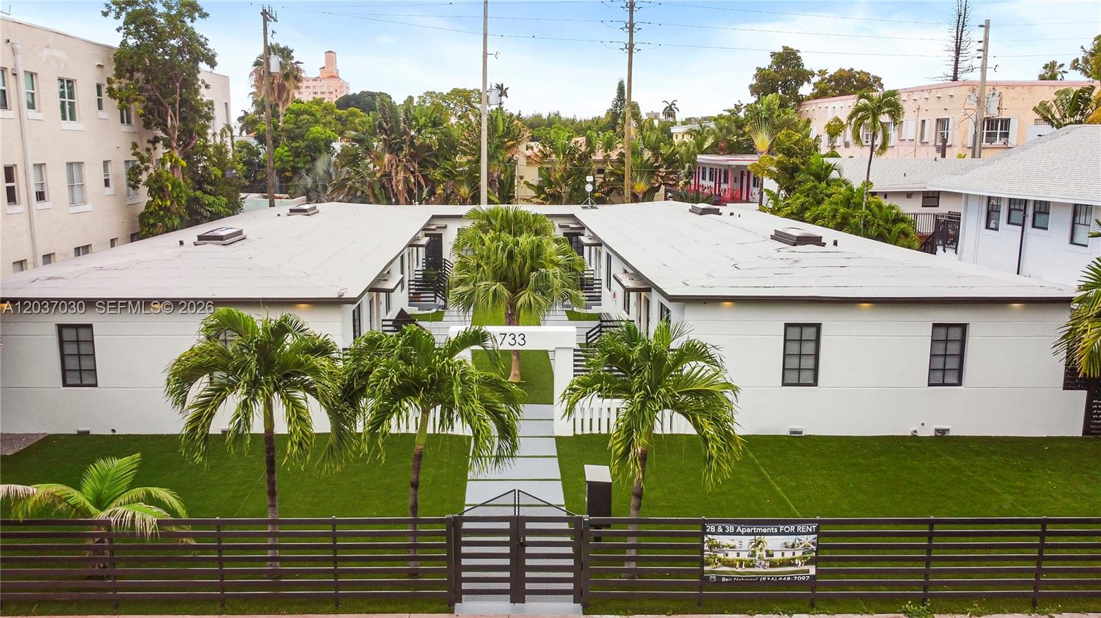 An exterior view of a white two-story apartment building with a central courtyard and palm trees — MIAMI BEACH, FL